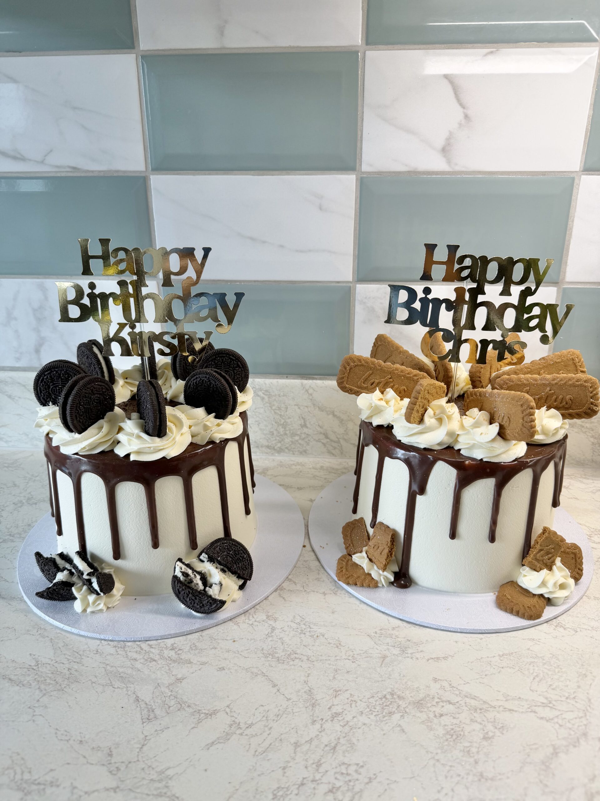 Two celebration cakes with white frosting and chocolate drip, topped with "Happy Birthday" toppers and cookies on each cake (oreo on left, biscuits on right).