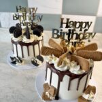 Two white birthday cakes with chocolate drip, piped cream, Oreo cookies, and cookie toppers reading 'Happy Birthday' at a marble counter.