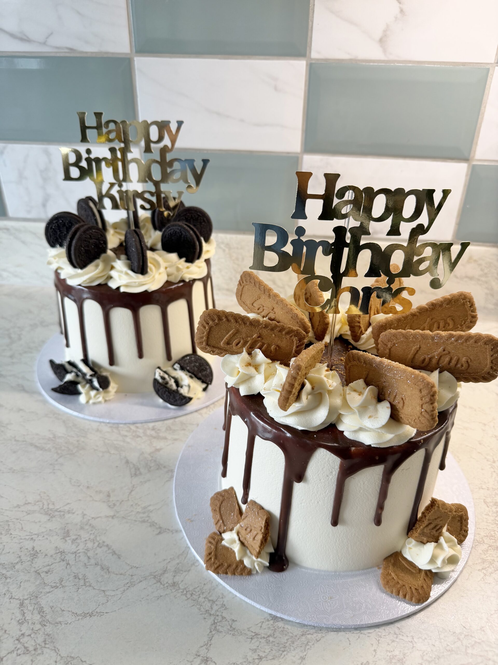 Two white birthday cakes with chocolate drip, piped cream, Oreo cookies, and cookie toppers reading 'Happy Birthday' at a marble counter.