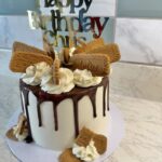 Birthday cake with white fondant, chocolate drip icing, and decorative whipped cream swirls topped by cookies and a 'Happy Birthday' topper.