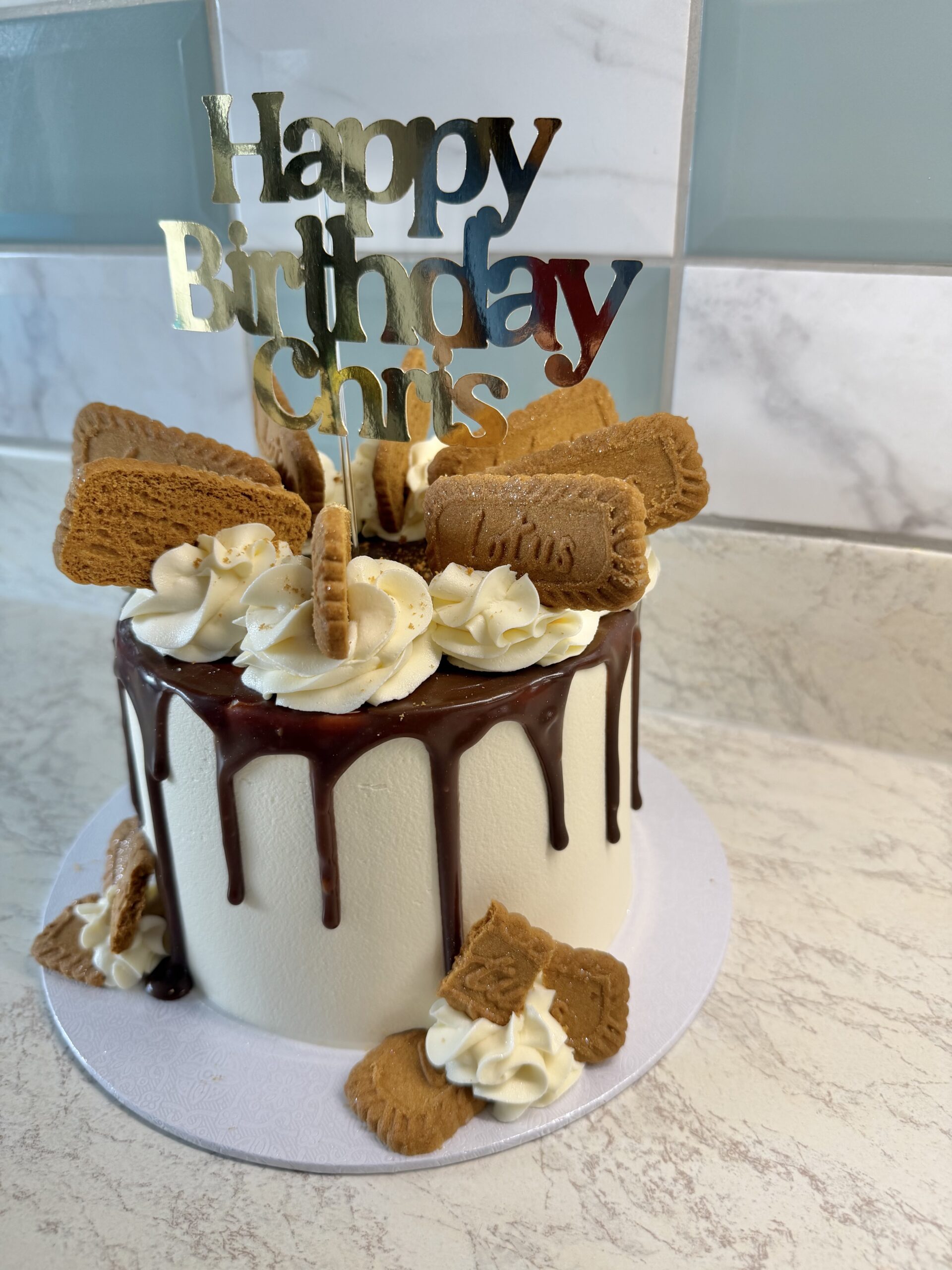 Birthday cake with white fondant, chocolate drip icing, and decorative whipped cream swirls topped by cookies and a 'Happy Birthday' topper.