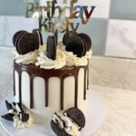 White fondant cake with chocolate ganache drip, vanilla buttercream swirls, and Oreo cookies on top; 'Happy Birthday' topper appears above the cake