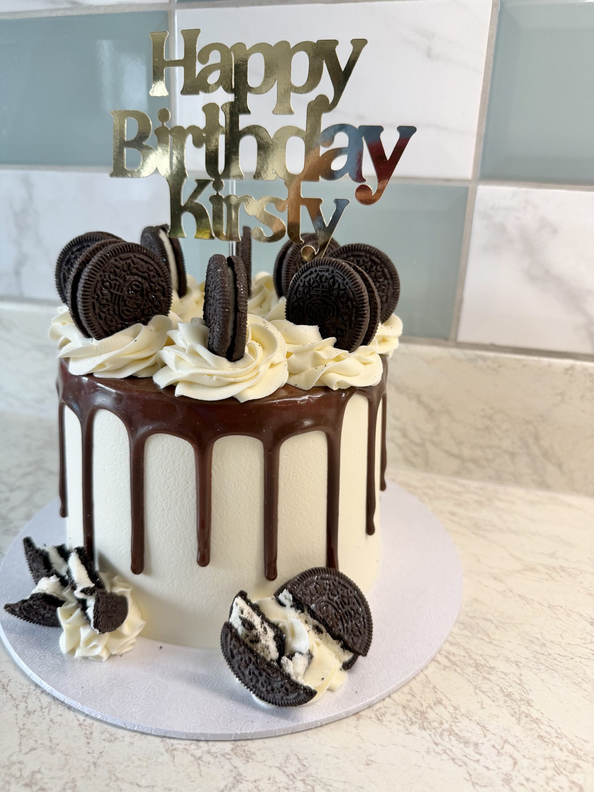 White fondant cake with chocolate ganache drip, vanilla buttercream swirls, and Oreo cookies on top; 'Happy Birthday' topper appears above the cake