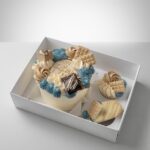 Boxed celebration cake adorned with blue and beige buttercream swirls, chocolate squares, and a small cake slice in a white gift box with cupcakes nearby