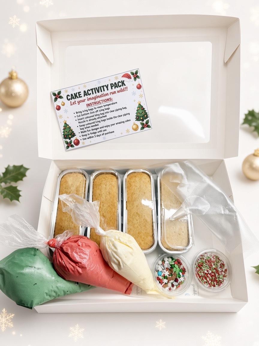 Open white gift box showing a Cake Activity Pack with four rectangular bake pans, piping bags in green, red, and white, plus sprinkle jars.